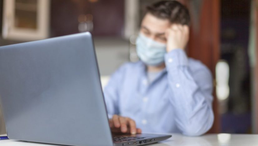 A man works from home on a laptop. Young man in protective respiratory mask against a virus. A businessman is sitting at a table in the kitchen. Coronavirus concept