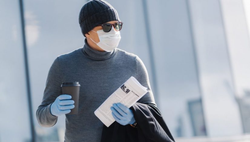 Outdoor image of male worker returns home from work, turns head aside, drinks coffee to go, holds newspaper, wears medical sterile mask and rubber gloves, prevents coronavirus spread. Infectious virus